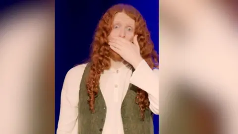 A man with long red hair and a green waistcoat clasps mouth over hands in shock.