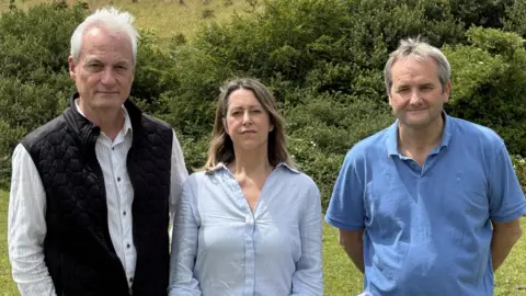 A woman with shoulder length brown hair wearing a blue shirt, standing between a man on her right with white hair, in a white striped shirt and black gilet, and a man with grey hair and a blue polo shirt on her left. They have some hedges and hills behind them.   