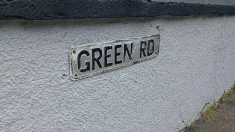A street sign reading 'green rd.' in capital letters. It is on a white wall.