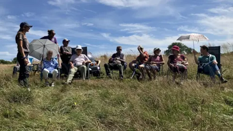 A row of elderly people sitting on camping chairs in the middle of a field. The grass has grown long and diverse. It is a clear day with wispy clouds, and most people are wearing hats and sunglasses or sitting under umbrellas to shield from the sun.