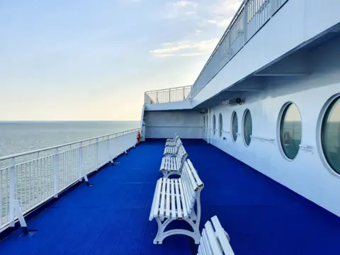 Getty Images An empty ferry Deck. there are benches and a view of the sea. the flooring is blue and the benches are white 