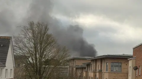 Simon Mayer The top part of blocks of two or three-storey flats and houses, with a thick dark plume of smoke behind them. There is also the top half of a tree.