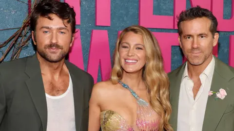 Getty Images Three people - Brandon Sklenar, Blake Lively and Ryan Reynolds - are dressed in formal attire and posing for a photo. They are stood in front of a large sign that reads "It Ends With Us"