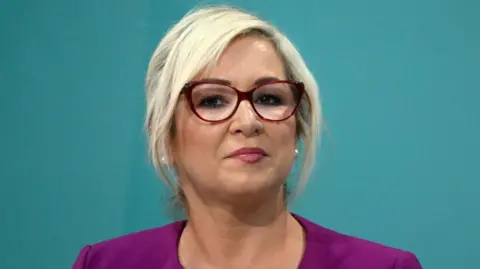 Sinn Fein vice president and Northern Ireland First Minister Michelle O'Neill is pictured before she gives her keynote speech at the Sinn Fein Ard Fheis on September 27, 2024 in Athlone, Ireland. She has blonde hair, which is up, and wears glasses. She has a plum coloured jacket on. 