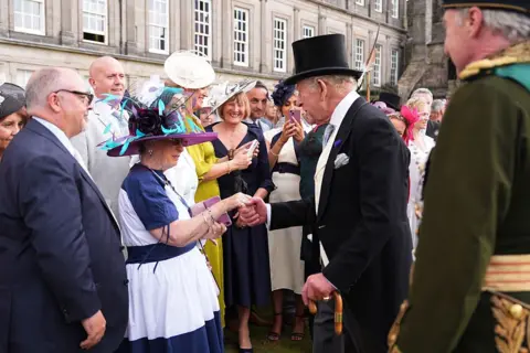 Getty Images A crowd of people outside Holyroodhouse meet the King. He is smiling and holding the hand of a woman in a formal dress. The King is in a morning suit and top hat.
