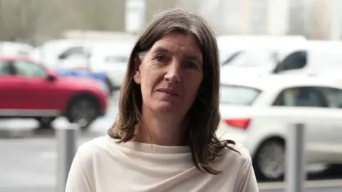 John Fairhall/BBC Rosie Pearson, a woman standing outside in a car park. She is looking directly at the camera and is wearing a cream blouse and her brunette hair is down. In the blurred background there are vehicles parked up.