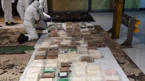 National Crime Agency Dozens of packages of drugs. They are lined up along the floor. Some contain white powder, others brown substances. Four people in all white hazmat suits are looking at the packages. 