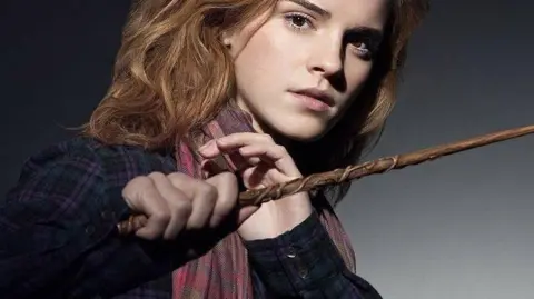 Warner Bros Hermione, with light brown wavy hair, wearing a plaid blue and purple jacket and pink scarf and holding a wooden wand out in front of her.