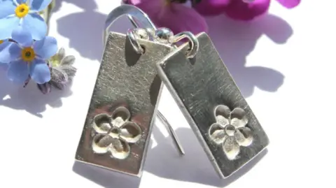 Emma Farley A pair of silver earrings which are rectangular in shape and have an imprinted five-petal flower design on them. Behind the earrings is a white background with blue and pink flowers.