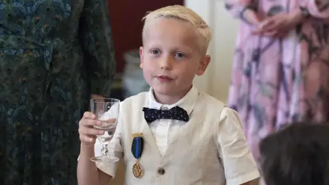 PA Media Rupert Williams with a glass of water. He wears a white short sleeved shirt, a beige waistcoat and a black bow tie. He has short blond hair. 