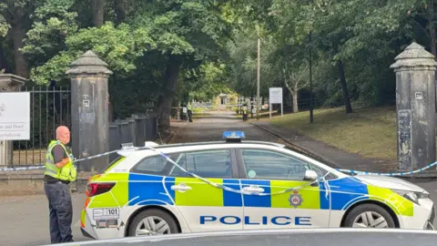 A police car and police tape outside a street lined with trees.