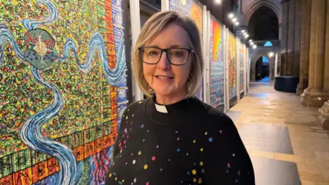 Reverend Eileen Wallis is wearing a black top with colourful dots on it and a clerical collar. She has large rectangular black-rimmed glasses and a blonde bob. A minster's passage can be seen behind her along with a colourful display alongside the passage depicting different biblical scenes.