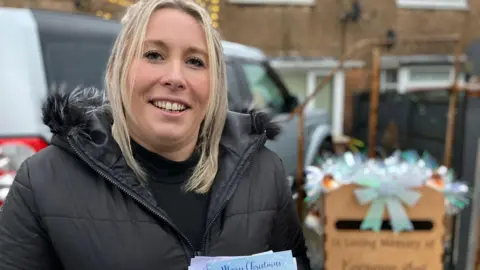 Amy Rogerson is pictured in front of the postbox, which is blurred. She is a white woman with shoulder-length blond hair. She is wearing a black puffer coat