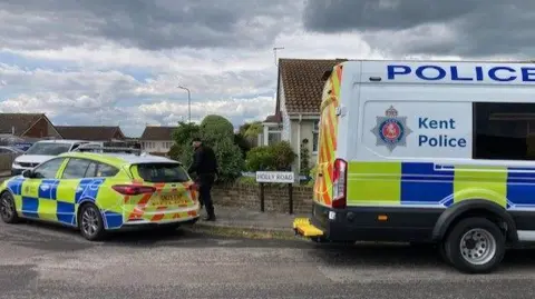 a police van and a police car park either side of a sign saying Holly Road, in front of a modern bungalow.