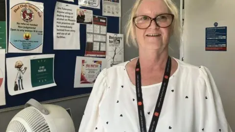 Somaiya Motara/BBC Sandra Skellern has a white top with blue haerts, blonde hair and glasses. She stands near a fan in front of a ntocie board.