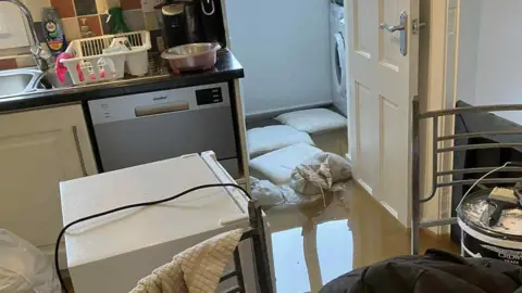 David Letherman A flooded kitchen. There is a fridge in the middle of the floor. Brownish water is covering the floor. There are sandbags at the door into the utility room, but they have not stopped the water coming in. 