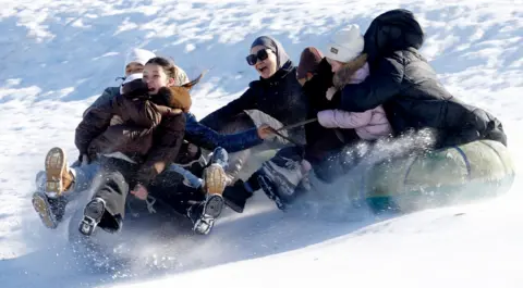 Igor Kovalenko/EPA/EFE A group of people laughing while going down a snowy slope on a sledge