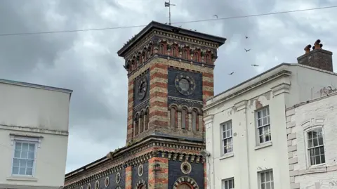 The New Market Buildings, which is of Italianate architecture, with pigeons flying around it's tower, which is topped with a weather vane