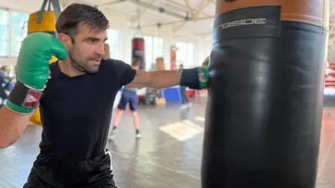 Chris hits a heavy punching bag hanging from the ceiling while people train behind him in the boxing club