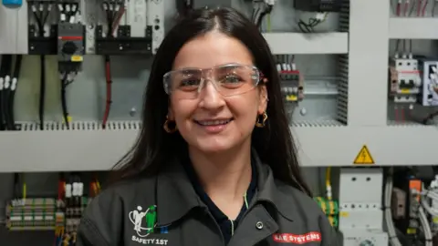 BAE Systems A woman with dark hair and wearing safety goggles and a black shirt with the BAE Systems branding on it. She's smiling at the camera and there's a wall of electrical equipment in soft focus behind her.