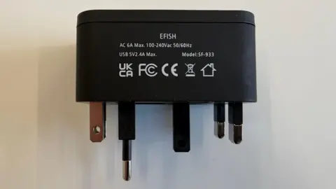 Electrical Safety First A universal travel adaptor, with information and safety markings displayed on its black plastic casing, is shown with all its pins retracted at once.