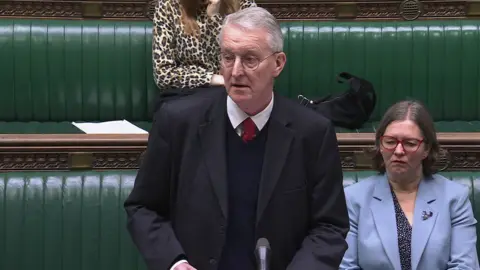 House of Commons Hilary Benn in black suit and jumper, wearing a white shirt and red tie, pictured in House of Commons with green benches in background.
A woman with grey hair, red glasses and a blue blazer is sat behind him