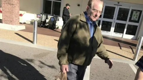 A man with thinning hair, glasses, a blue shirt, and a green fleece walks across a car park. He looks down at the ground