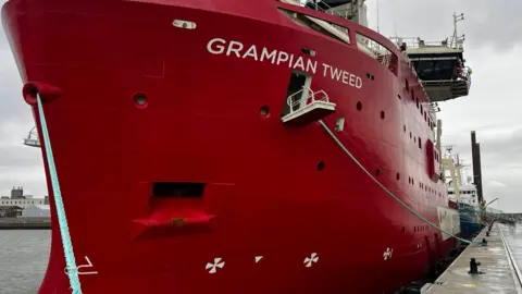 John Fairhall/BBC Grampian Tweed is a large red ship seen docked at a port. A white gang plank is open halfway down the side of the ship. 