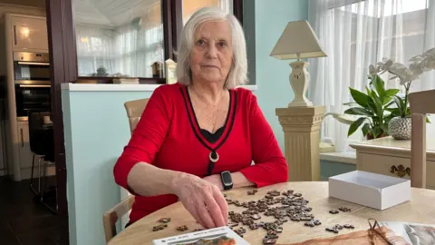 Dorothy sits at a table in her conservatory. She is doing a jigsaw and looking into the camera.