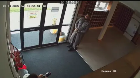 CCTV view of a man in a dark grey outfit smiling by the door of a hallway. Another man sits in the corner putting on a pair of shoes.