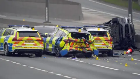 PA Media Three police cars and a police van in the middle of the empty A1. One looks damaged from the back. A normal car is on its side next to the central reservation.