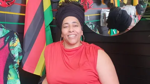 Woman with black headwrap and red t-shirt smiling in front of Caribbean flags and mirror behind reflecting shop display.