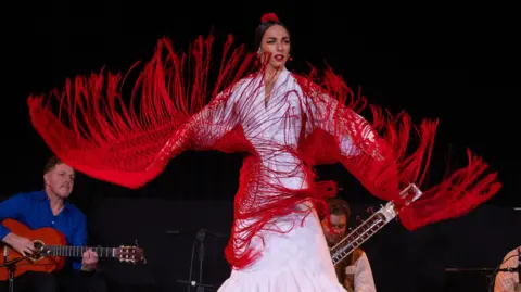 Flamenco artist wearing a long white dress and read shawl with fringes on it. There are musicians behind her. The dancer is mid movement. 