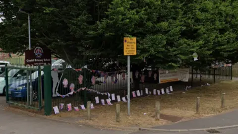 The photo has been taken from the street outside a primary school. There is a green fence just off the footpath, which has union jack bunting hung on it. To the left is a brown sign which reads 'Goxhill Primary School.' The are multiple cars parked in a car park behind it. 