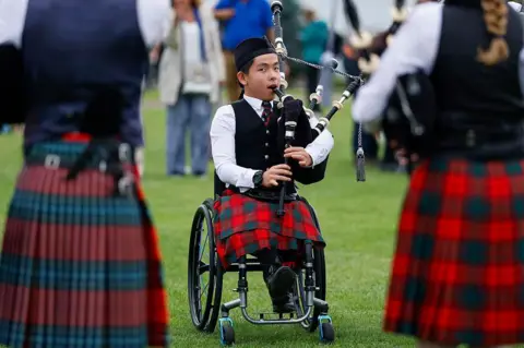 Getty Images Young person in a wheelchair playing a set of bagpipes. They are wearing a  traditional piper outfit with hat