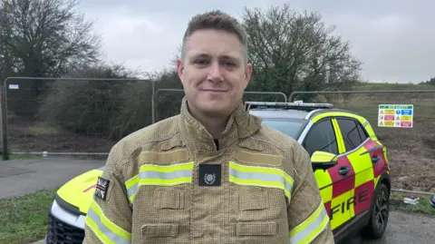 Kate Bradbrook/BBC A firefighter wearing protective gear with reflective yellow stripes stands outdoors in front of a marked fire service vehicle. 