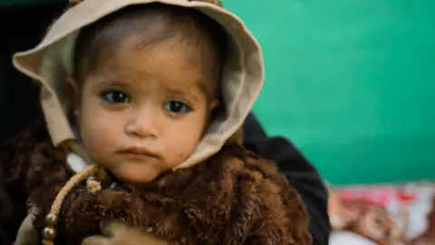 A young child wearing a thick brown top and a beige hood around their head looks at the camera