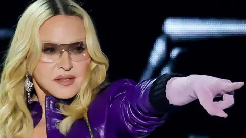 Clad entirely in purple and wearing sunglasses, with long, blonde hair framing her face, Madonna points into the distance as she performs on the main stage at Coachella festival