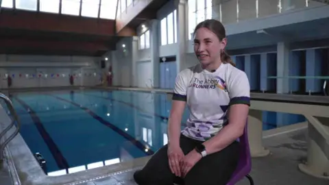 Carys Westcott, smiling at the camera, sitting on a purple chair. She is wearing her hair up and has a white t-shirt on and black trousers.