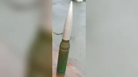 A military shell about 45cm tall sits on an outdoor surface. Its propellant section is brass coloured, with a large silver projectile on top. 