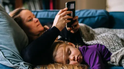 Getty Images A young girl in a purple sweatshirt lies next to a woman in a black jumper on a couch. Both are covered in blankets 