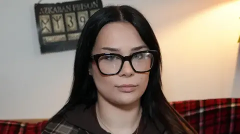 Poppy Stopforth slightly smiles at the camera. She has long dark hair, glasses and wears a grey and black chequered jacket. 