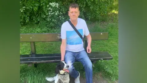 Steven Mead wearing a white T-shirt with a light blue square at the front of it and blue jeans. He is sitting on a park bench and smiling at the camera.
He has a dog sitting next to him.