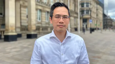 A man in a white shirt stands in a city square outside Birmingham Council House, which is a sandstone building with pillars. Yip has dark greying hair brushed back and wears rimless glasses.