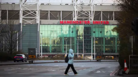 A woman in a blue rain coat with a black rucksack crosses a road, with the Old Trafford stadium seen towering behind her. 