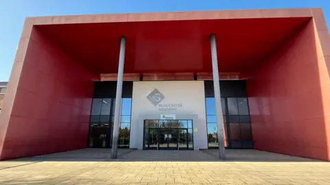 Image of the front of Gloucester Academy. It has a paved area at the front and the building has large glass windows surrounded by red walls.