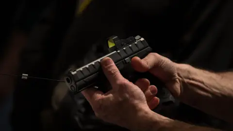 A pair of male hands holding a black Beretta pistol against a shadowy backdrop.