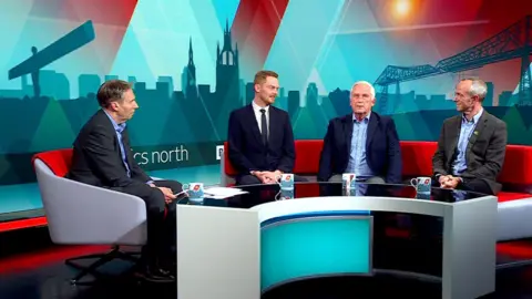 Presenter Richard Moss sitting on a chair in the Politics North studio. He is looking to his left at Luke Myer, Glen Sanderson and David Francis who are sitting on a curved red sofa. Coffee mugs sit on a curved table is in front of them.