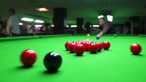 Red and black snooker balls are in motion on the green table. There is someone pulling the cue in the background but they are out of focus.