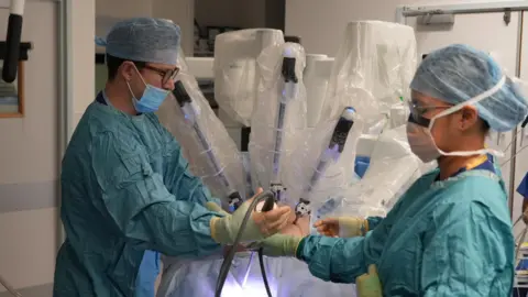 Shaun Whitmore/BBC Surgeon Nicholas Penney adjusts the camera for the surgical robot in one of the theatres of the Norfolk and Norwich Hospital. His right hand has a camera in his hand, while another member of theatre staff supports the cable for the camera. They are both dressed in green theatre gowns, hair nets and face masks